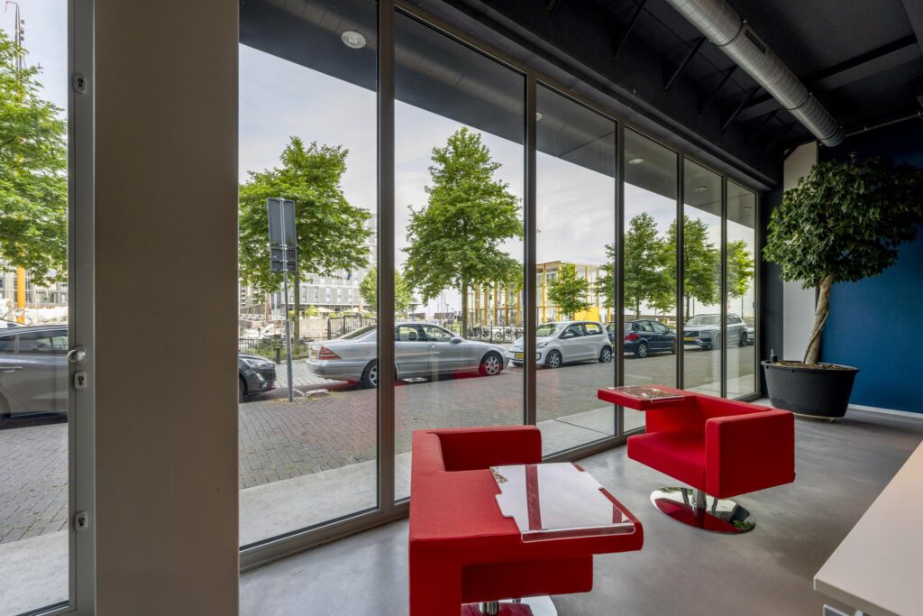 Modern waiting area with red chairs and large windows overlooking Krijn Taconiskade street and parked cars.